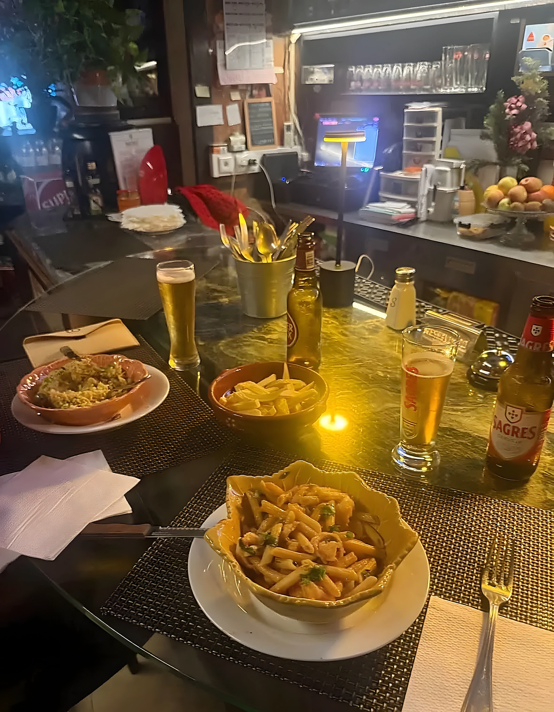 Massa com gambas em prato de barro, outra travessa de arroz, batatas fritas e cerveja no balcão da taberna.